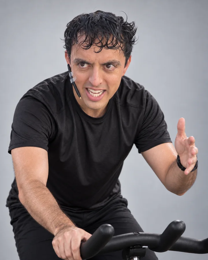José Ramones como coach de spinning, vestido de negro sobre una bicicleta estática